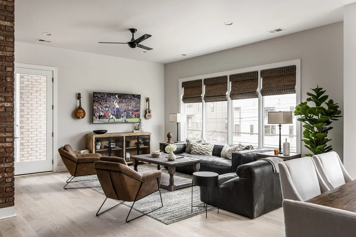 A spacious living room featuring a large sectional sofa, flat-screen TV, wooden coffee table, an abundance of natural light, and indoor plants. The room is designed for comfort and group activities, ideal for streaming playlists or relaxing after a day out.