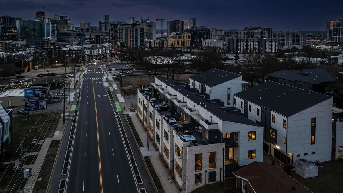 An exterior scene showcasing modern townhouses with the illuminated city skyline in the background and a street with bike lanes in the foreground. The mood is contemporary and vibrant, suggesting evening strolls, city exploration, and an active urban lifestyle.