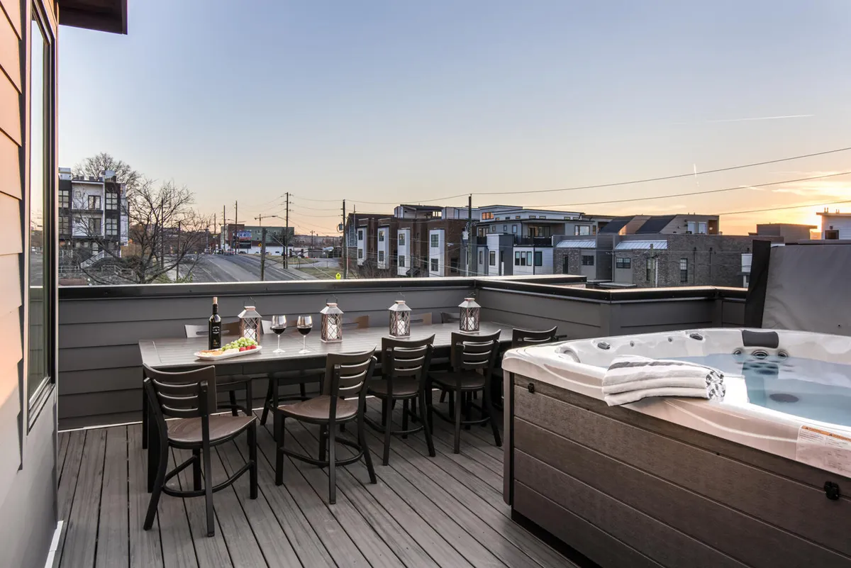 An inviting rooftop patio features a dining table with chairs, a bubbling hot tub, wine glasses, a bottle of wine, and lanterns set for evening ambiance. The city skyline is visible in the background, hinting at elevated views and a lively social atmosphere perfect for sunset gatherings.