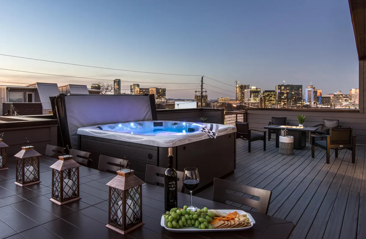 The image showcases an inviting rooftop hot tub lit with vibrant LED lights, surrounded by stylish outdoor dining complete with wine and snacks. Lanterns cast a warm glow as the sun sets over the Nashville city skyline, creating a luxurious and tranquil setting ideal for socializing, relaxing, or enjoying a romantic evening.