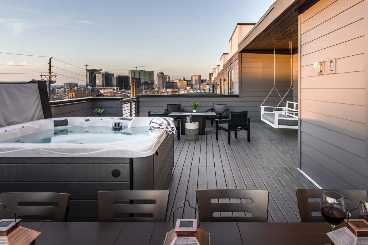 This image showcases a rooftop deck with a bubbling hot tub, an inviting outdoor seating area, a dining table perfectly set for al fresco meals, and sweeping panoramic views of the Nashville city skyline at sunset. The scene is tranquil, highlighting people relaxing in the hot tub and engaging in social conversation, with city lights beginning to twinkle in the background.