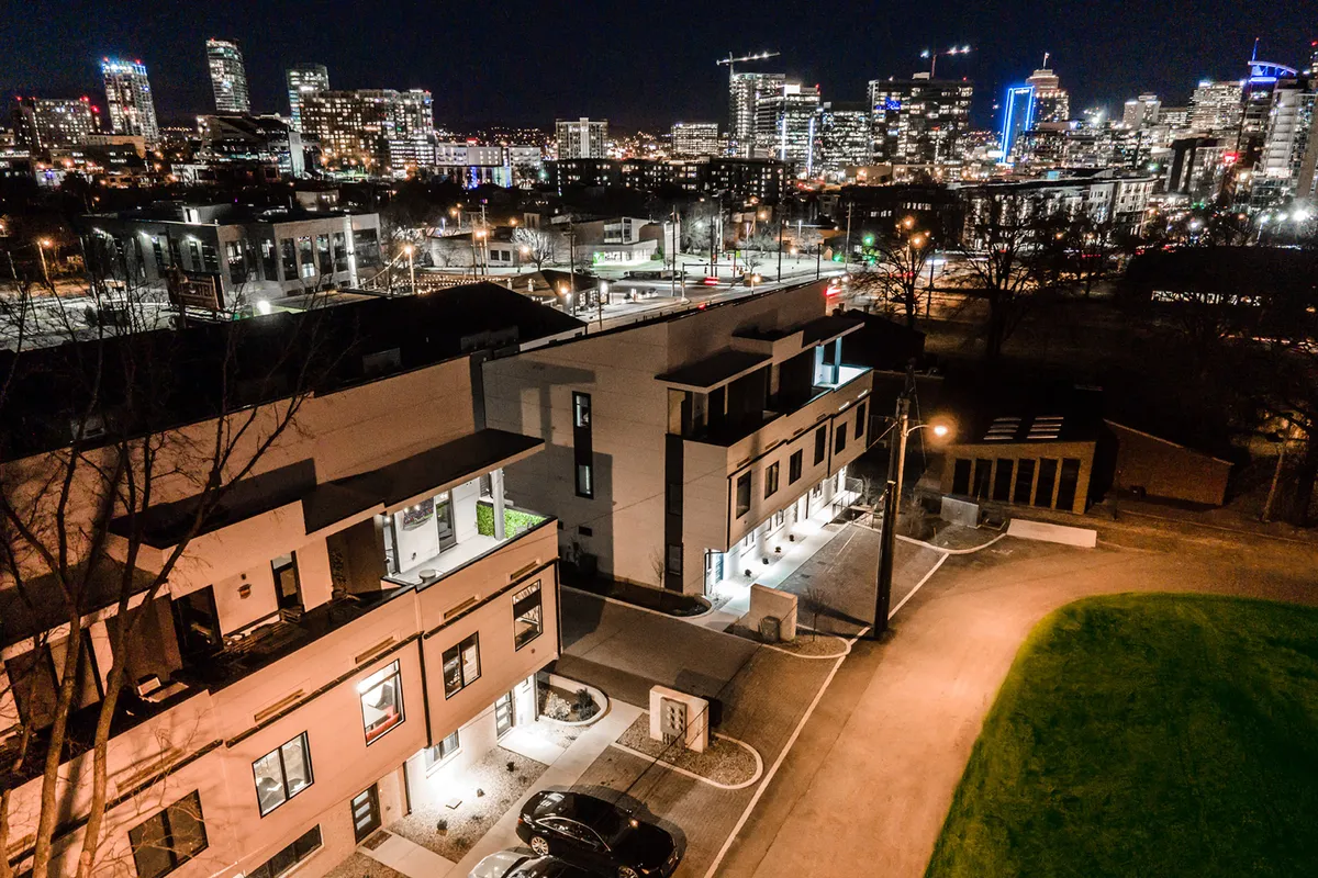 This exterior shot highlights the modern architecture of urban townhouses set against the city skyline, with street lights and green space visible. The image captures the vibrancy of city life, suggesting proximity to major business centers and an active, dynamic environment.