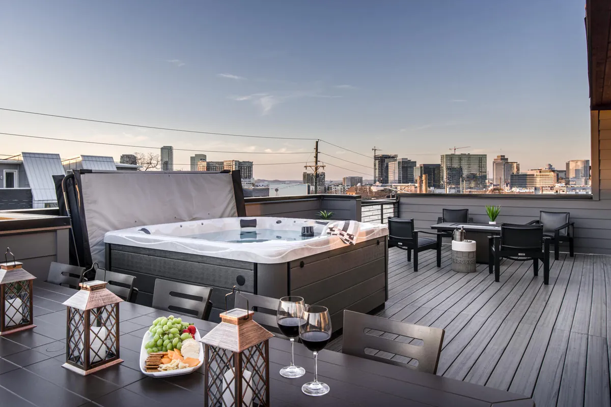 A rooftop retreat featuring a hot tub, elegant dining table, lanterns, wine glasses, and a panoramic view of the city skyline. The setting is luxurious and intimate, ideal for relaxing after a night out in the city.