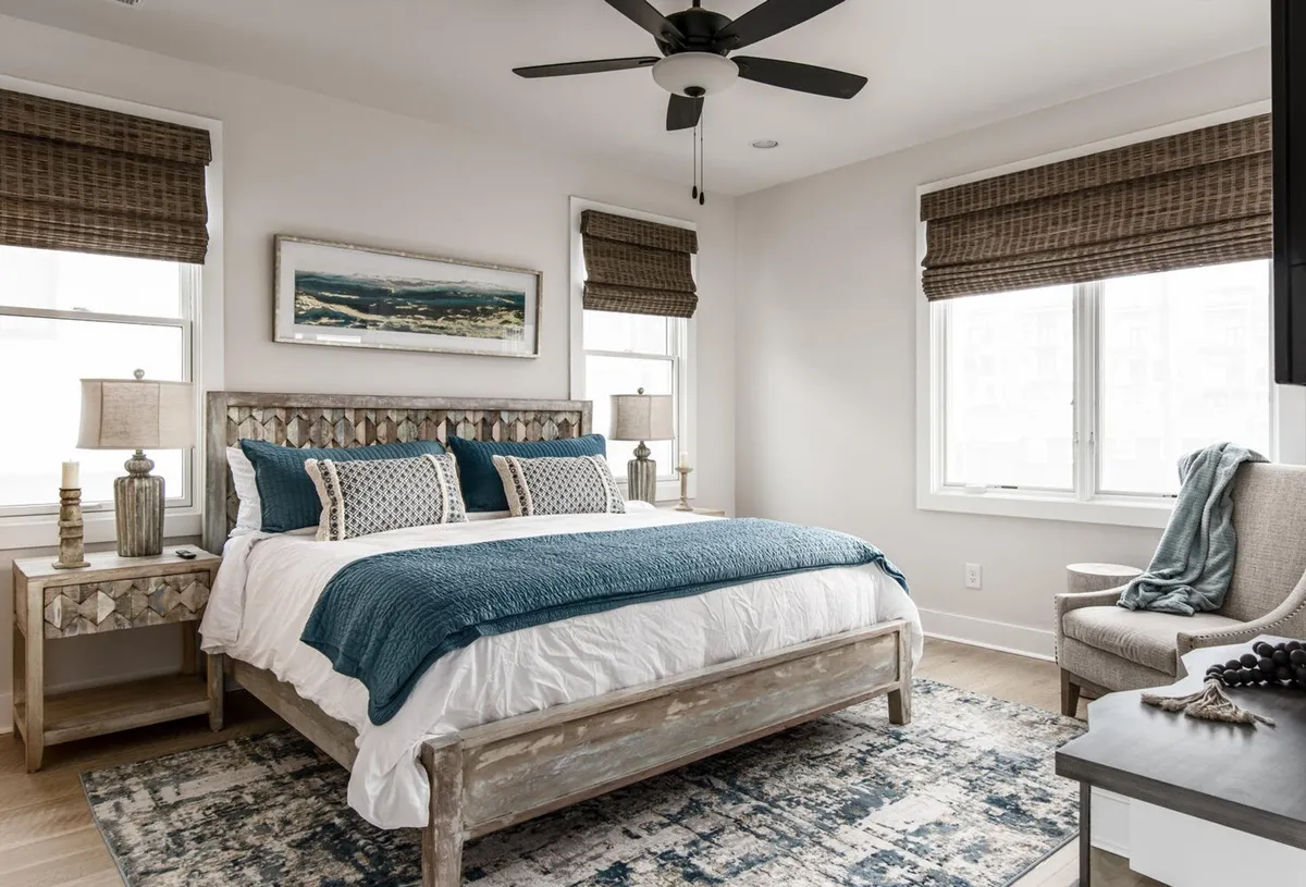 A serene bedroom featuring a bed with textured bedding, nightstands with lamps, tasteful artwork on the wall, windows with blinds letting in natural light, and a comfortable reading chair in the corner.